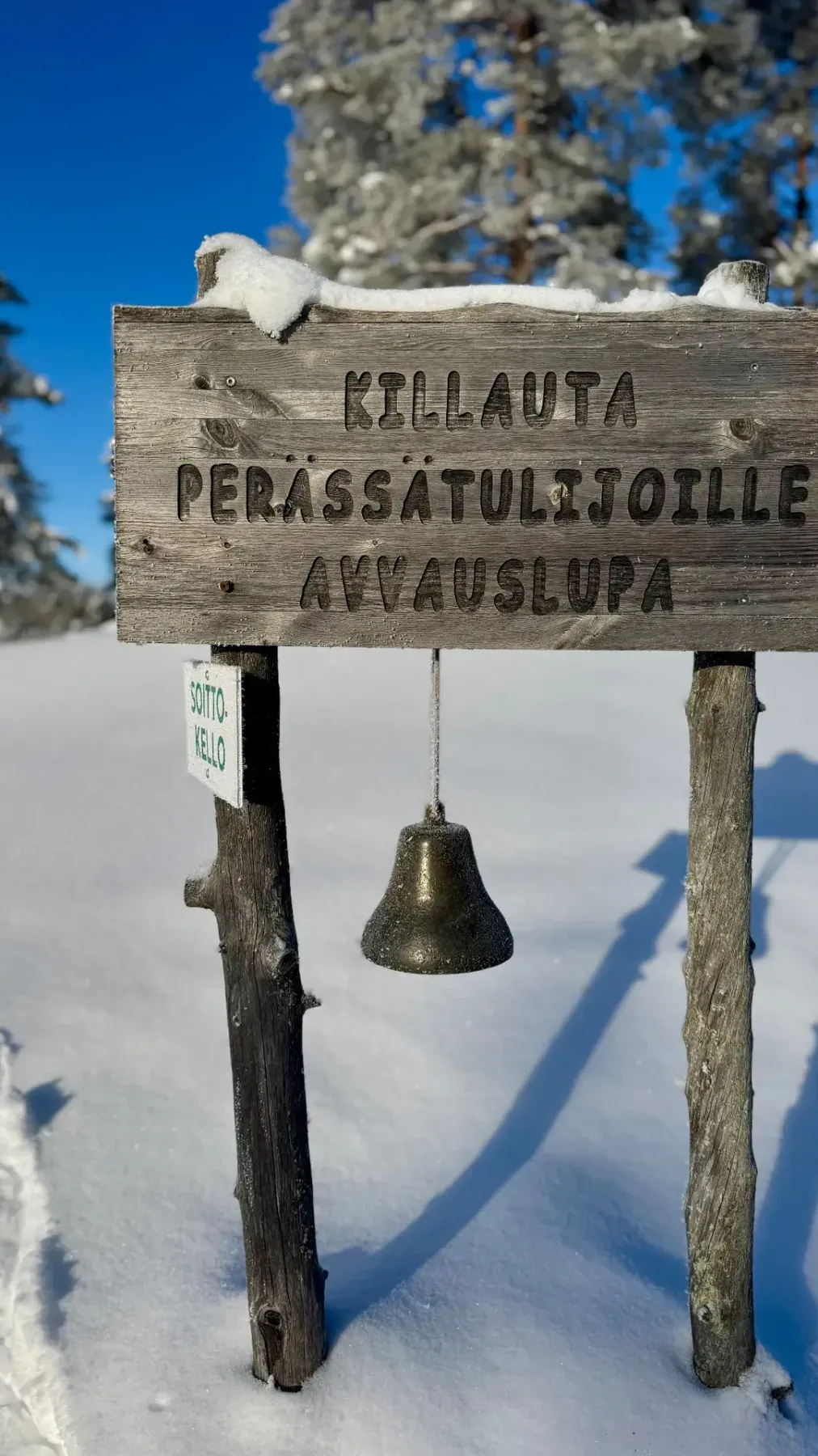 Finnisches Holzschild mit Glocke am verschneiten Wegesrand vor blauem Himmel