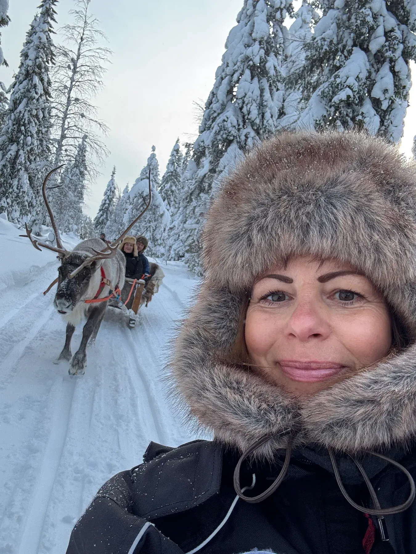 Selfie mit Fellkapuze, im Hintergrund ein Rentiergespann auf dem verschneiten Weg