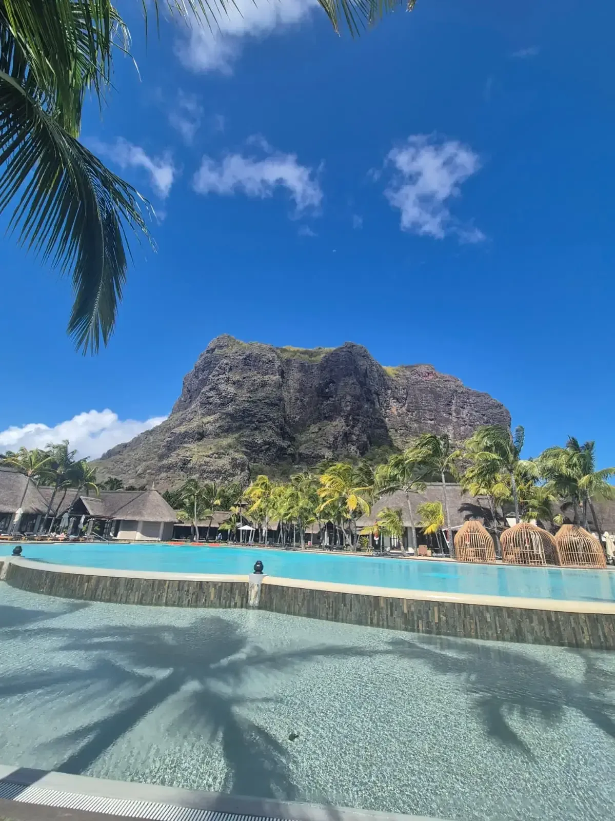 Hotelpool mit Palmenschatten, dahinter strohgedeckte Bungalows und ein mächtiger Felsberg unter blauem Himmel